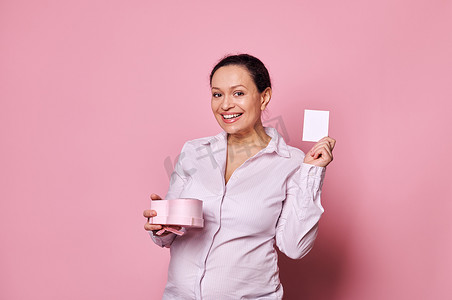 怀孕的中年漂亮女人对着镜头微笑，同时展示粉红色背景中突显的贺卡和礼盒