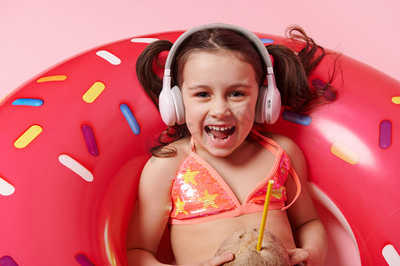 淘气摄影照片_带耳机和可可饮料的滑稽淘气小女孩的特写，在甜甜圈游泳圈内，粉红色背景