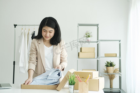 t恤女摄影照片_亚洲女服装店老板折叠一件 T 恤，然后用纸板包裹箱包装。