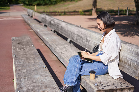 亚洲女孩的肖像坐在公园的长椅上，用笔在她的数字平板电脑上画画，开心地笑着，变得有创意