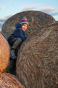 一个淘气的小男孩在秋天的傍晚在夕阳的照耀下爬到一捆稻草的顶部