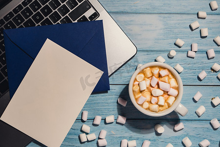 空的问候语或邀请卡模拟蓝色信封，白色咖啡杯和棉花糖笔记本电脑键盘放在蓝色木制桌子上。