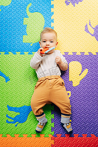 身穿白色紧身连衣裤和棕色裤子的小婴儿躺在彩色地毯上，抱着并啃着一个明亮的拨浪鼓。