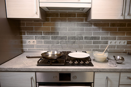 玻璃碗中的木勺，配煎饼面糊，厨房台面上的不锈钢厨房用具，黑色炉子上的白盘和煎锅。