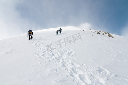 登山者背着背包，沿着白雪皑皑的山脊向上走。