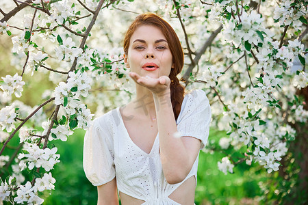 一个穿着浅色连衣裙的可爱女孩的肖像，背景是一棵盛开的白树，将鲜花从她的手掌吹进相机