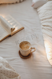 装饰组合摄影照片_舒适的晨间组合物，配有咖啡杯和一本书，书页在床上折叠成心形