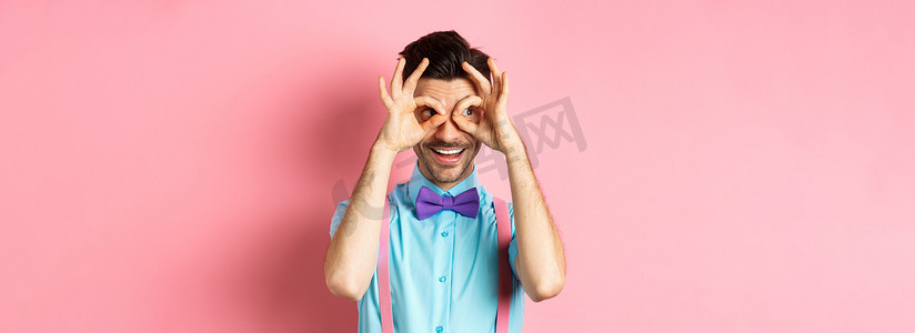戴着领结和吊带的快乐男人透过手持双筒望远镜看着，微笑着，看到有趣的假期优惠，盯着粉红色背景中的相机