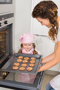 烤箱饼干摄影照片_妈妈在小女孩的注视下从烤箱里拿出饼干