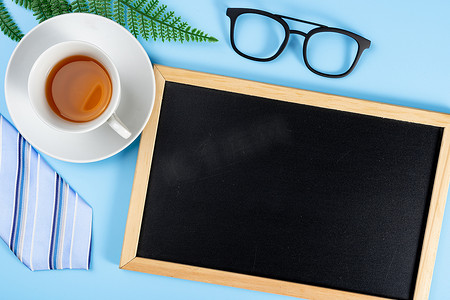 快乐的父亲节背景概念与蓝色领带眼镜、叶子、茶杯和蓝色背景的黑板，带有文本复制空间。