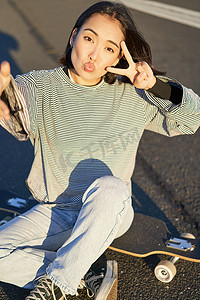 坐在滑板上的女孩摄影照片_亚洲女孩坐在滑板上的自拍，在智能手机上拍照，微笑着展示和平 V 字牌