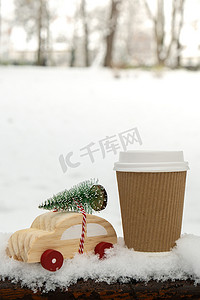 木制汽车载着圣诞树，纸杯在雪地上模拟咖啡或热巧克力。