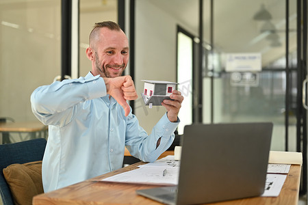 白人男性房地产经纪人拿着房子模型在通过笔记本电脑与客户进行视频通话时显示大拇指朝下