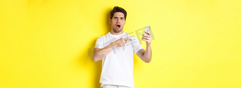 懂生活摄影照片_困惑的人用手指指着手机屏幕，站在黄色背景上，听不懂