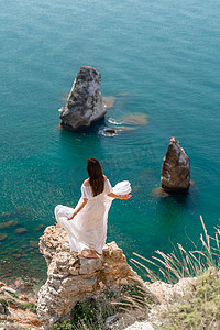 在蓝天大海的映衬下，一位身穿白色轻裙长腿的美丽少妇挥舞着白色长裙站在海面之上的悬崖边上。