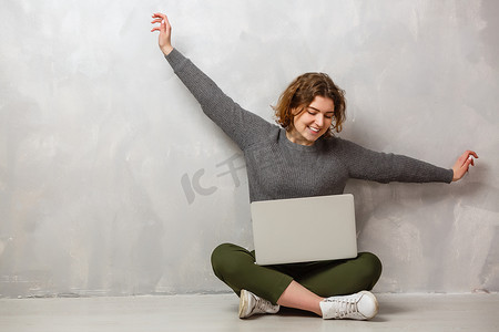 学生看电影摄影照片_满意的女性画像，带着美丽的微笑，享受着用银色电脑看电影，坐在灰色墙壁上的地板上的莲花姿势
