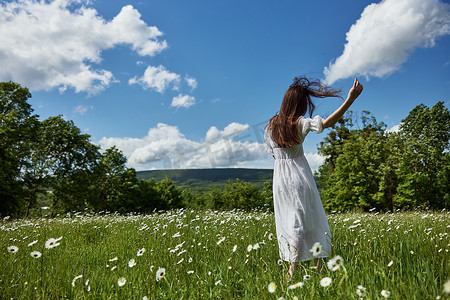 一个长着红色长发的女人背对着镜头站着，身着浅色连衣裙站在蓝天映衬下的雏菊花田里，望着远方