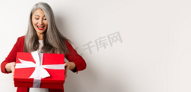 头发灰白的快乐老妇人收到礼物，看着红色礼盒，惊讶地笑着，站在白色背景上