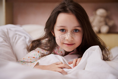 特写一个可爱的小女孩，看着相机，躺在床上，上面铺着舒适柔软的白色毯子