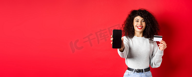 展示塑料信用卡和空智能手机屏幕的快乐漂亮女孩，展示账户或应用程序，站在红色背景上
