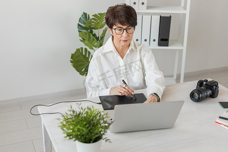 中年女性平面设计师或摄影师在现代办公室工作时使用数字图形平板电脑，专业女性修饰师坐在现代工作区，通用设计平板电脑和附近的相机