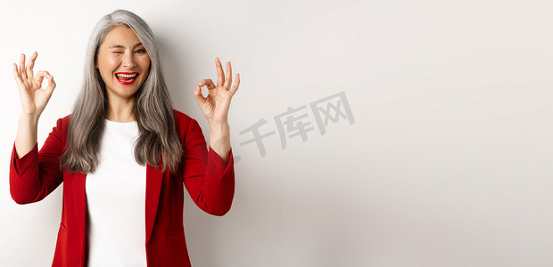 保证手势摄影照片_站在白色背景下，亚洲职业女商人表现出 OK 的手势，眨眼，高兴地微笑，保证或推荐某事