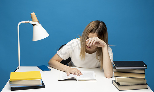 穿着白色休闲服的疲惫的年轻女学生坐在桌旁，在大学或学院图书馆看书。