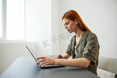红头发的女人坐在办公室靠窗的桌子旁全职工作，在笔记本电脑上打字