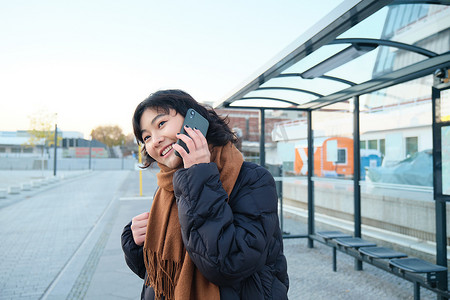 美丽的微笑韩国女孩，在公共汽车站等车，使用公共交通工具，用手机聊天，去城市的某个地方