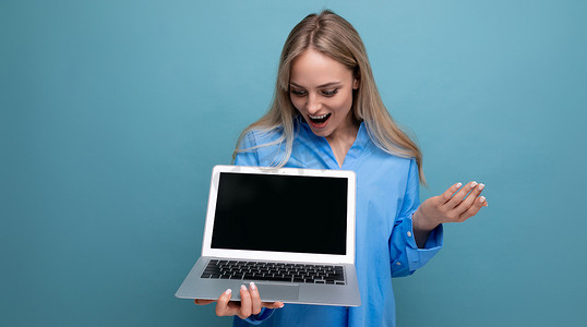 惊喜发现摄影照片_快乐幸运的金发女孩惊喜地发现，她手里拿着一台笔记本电脑，蓝色背景上有一个空白的网页空间