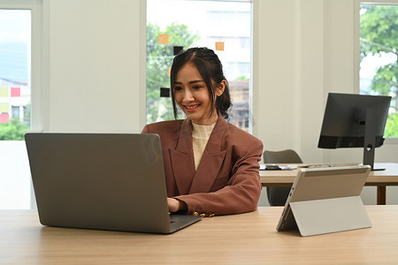 女性电脑摄影照片_在现代办公室的木桌上使用笔记本电脑和数字平板电脑的职业女性经济学家
