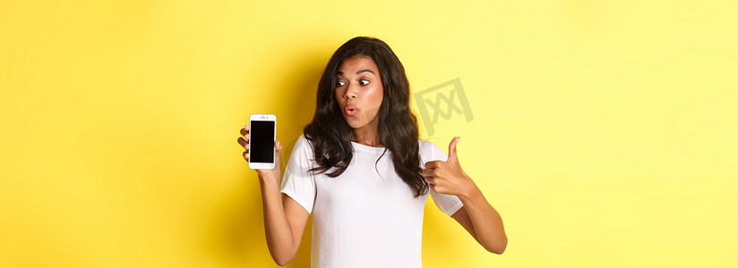 漂亮的非洲裔美国女模特肖像，推荐一款应用，展示手机，看起来很兴奋，站在黄色背景上