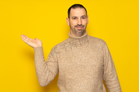 黄色促销摄影照片_40 多岁的留着胡子的西班牙裔男子身穿米色高领毛衣，在广告概念中展示一种虚构的产品，带有复制空间，与黄色工作室背景隔离。