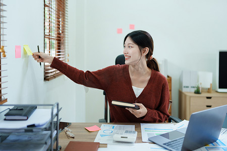 财务报表分析报告摄影照片_一位体贴的亚洲女商人的肖像，她正在查看财务报表并使用办公桌上的电脑和笔记本制定营销计划