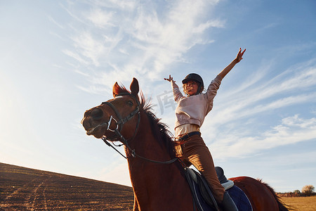 防护帽摄影照片_阳光明媚的白天，戴着防护帽的年轻女子和她的马在农田里