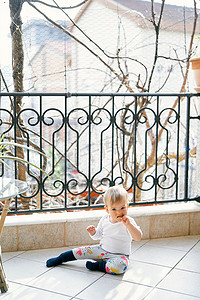 穿着白色衬衫和亮色裤子的小孩坐在靠近桌子的阳台上，在树的背景下啃着饼干