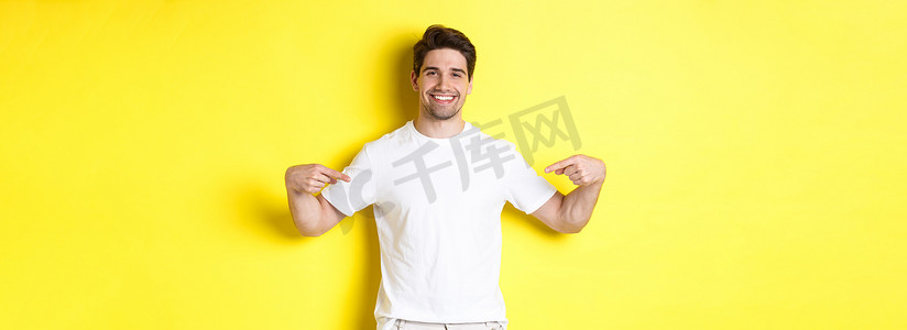 快乐的帅哥用手指指着你的标志，在他的 T 恤上展示促销，站在黄色背景上
