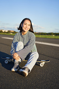 春天户外运动女摄影照片_亚洲女滑手的垂直镜头，坐在她的滑板上微笑，享受阳光明媚的日子，在户外空荡荡的路上在长板上巡航