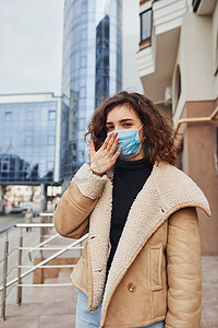 隔离期间，戴着防护面罩的卷发小女孩在商业大楼附近咳嗽的画像。