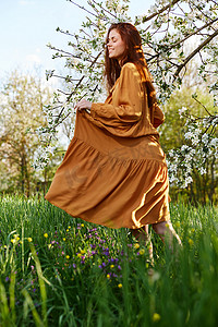 绿色树苗条摄影照片_一个快乐、苗条、甜美的女人穿着橙色长裙站在一棵开花的树旁高高的草丛中，开心地微笑着掀起裙子的下摆