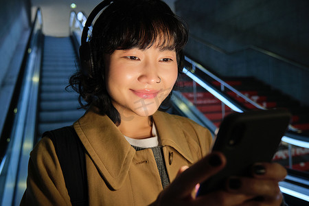 戴着耳机的可爱亚洲女孩的特写，在上下班途中选歌听，下自动扶梯去地铁，微笑着在手机上阅读信息