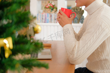 男人拿着咖啡摄影照片_男人拿着咖啡杯 & 在圣诞节期间在家里使用电脑。
