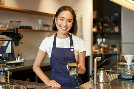 亚洲咖啡师女孩在柜台的画像，向想要非接触式支付、点菜、站在咖啡馆的客户展示卡片机