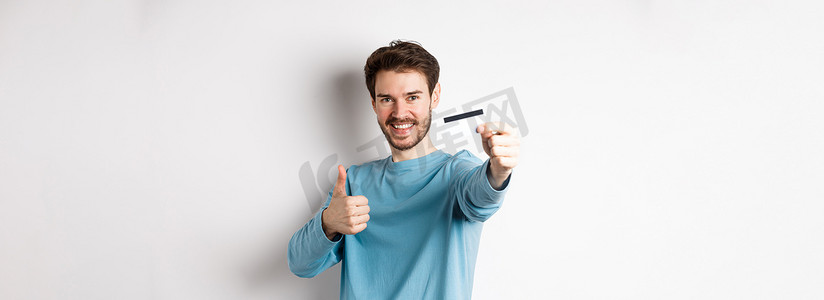 报价摄影照片_英俊的白人男子站在白色背景上，用大拇指展示塑料信用卡，推荐并称赞良好的银行报价。