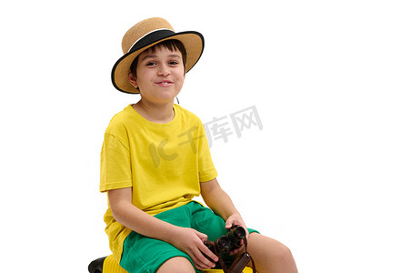 微笑的旅行男孩，戴着草帽和黄绿色的夏装，拿着双筒望远镜，对着白色的相机微笑
