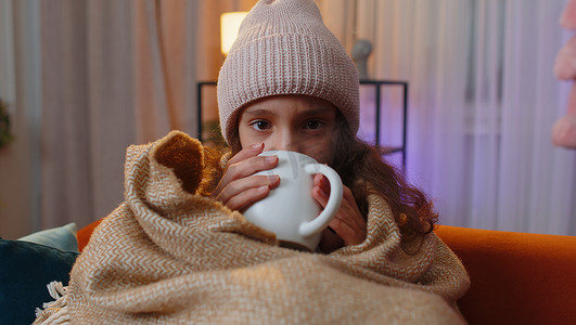 生病的青春期前女孩戴着裹着格子花呢的帽子，独自坐在沙发上喝着热茶，冷得瑟瑟发抖
