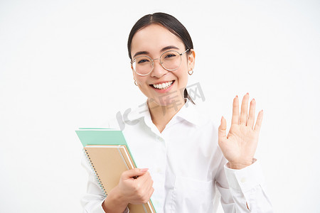友好的日本学生形象，女孩挥手，打招呼，戴眼镜，拿着笔记本，站在白色背景上