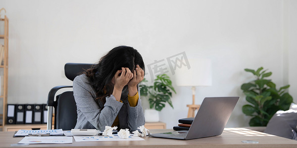 员工电脑摄影照片_压力大的疲惫女商人坐在家里的桌子旁，用笔记本电脑工作，手握着太阳穴，偏头痛发作。