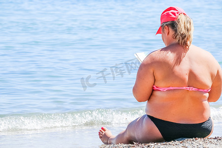 一个戴着帽子的女人，坐在沙滩上，看书，凝视着大海。超重的女性身体在海滩上。加大码的女人。