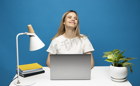 身穿休闲白衬衫的年轻成功快乐的员工女商人坐在办公桌前工作，电脑笔记本电脑工作区在蓝色背景工作室做孤立的赢家手势。